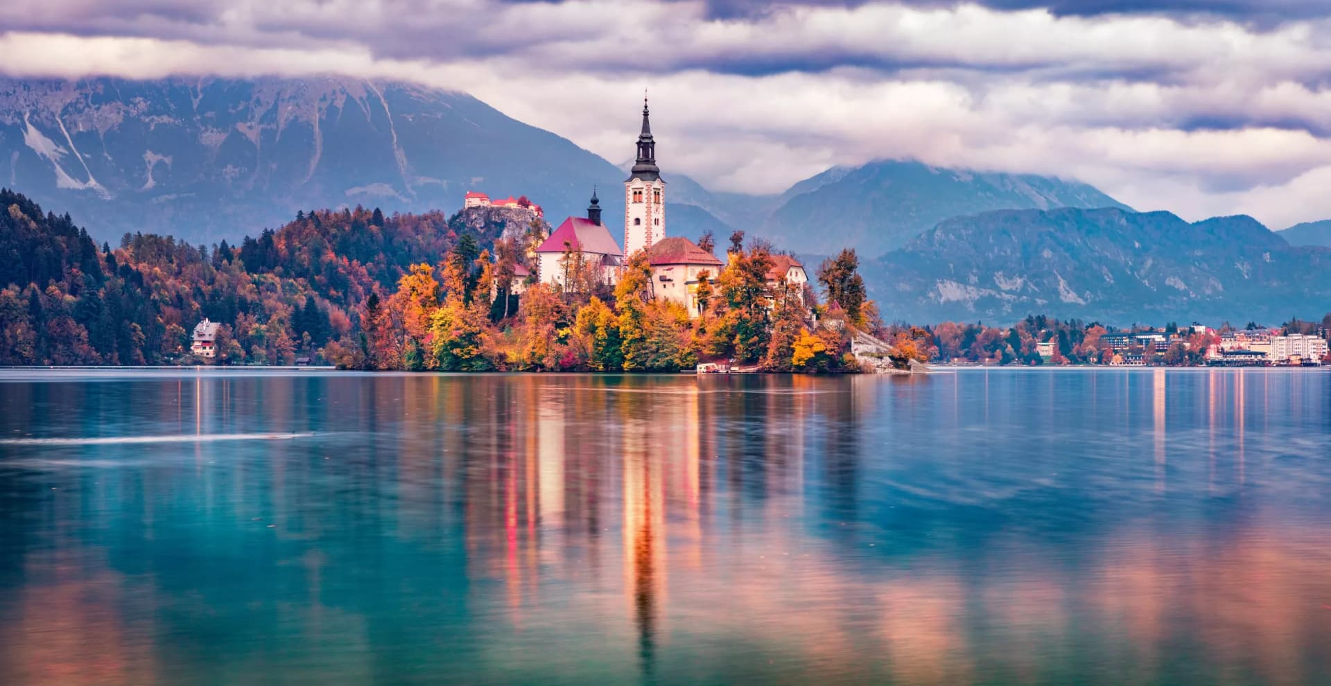 Church on island in Lake Bled with autumn trees and mountains under cloudy sky