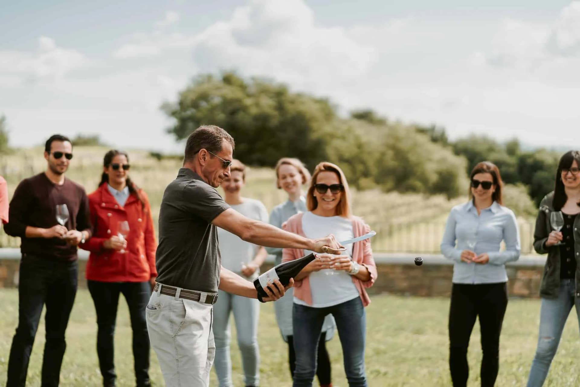 Wine tasting group watches sabrage outdoors with vineyards in background, Goriska Brda.
