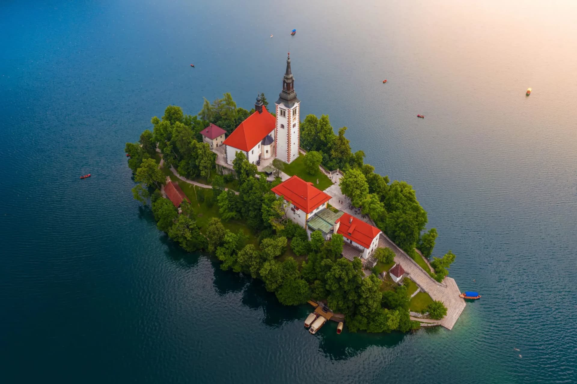 Aerial view of Bled Island church with red roofs surrounded by green trees on blue lake water.