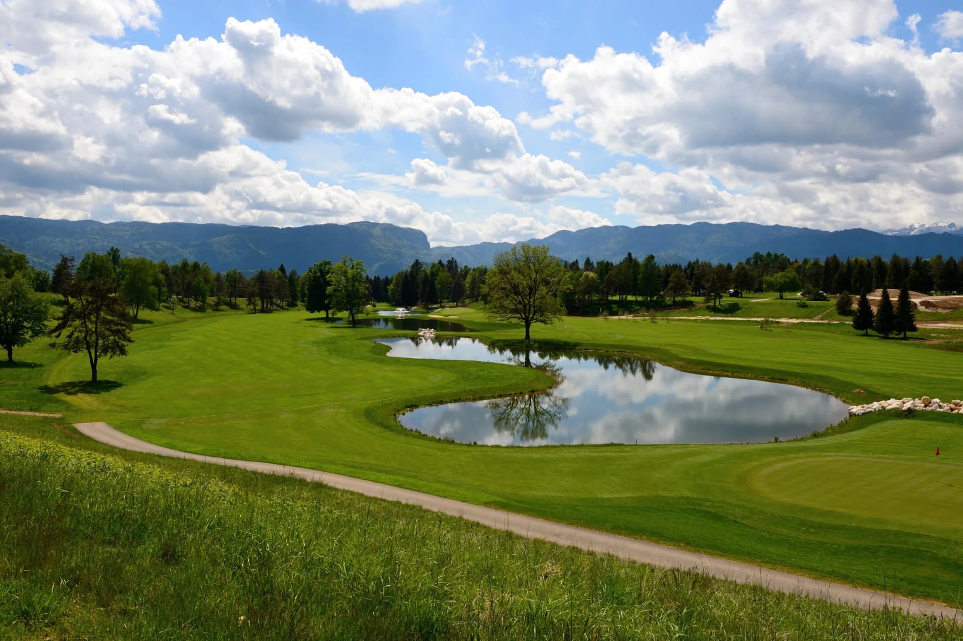 Golf course with pond reflecting clouds, set against forested mountains under a blue sky.