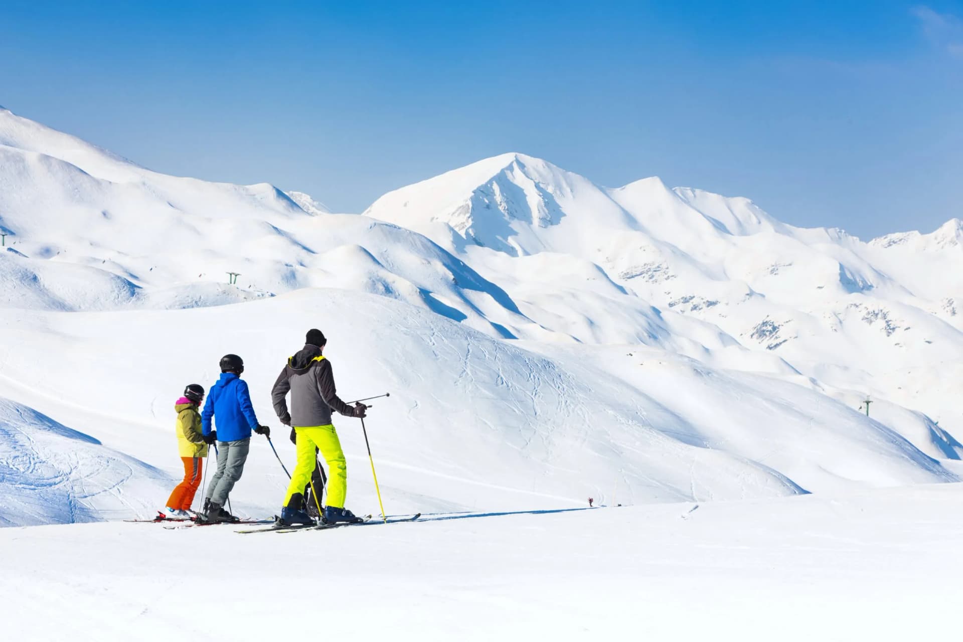 Skiers standing on a snowy slope with massive snow-covered mountains under a clear blue sky.