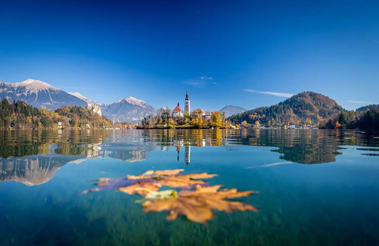 Lake Bled island church with autumn foliage, mountains, and floating maple leaf.