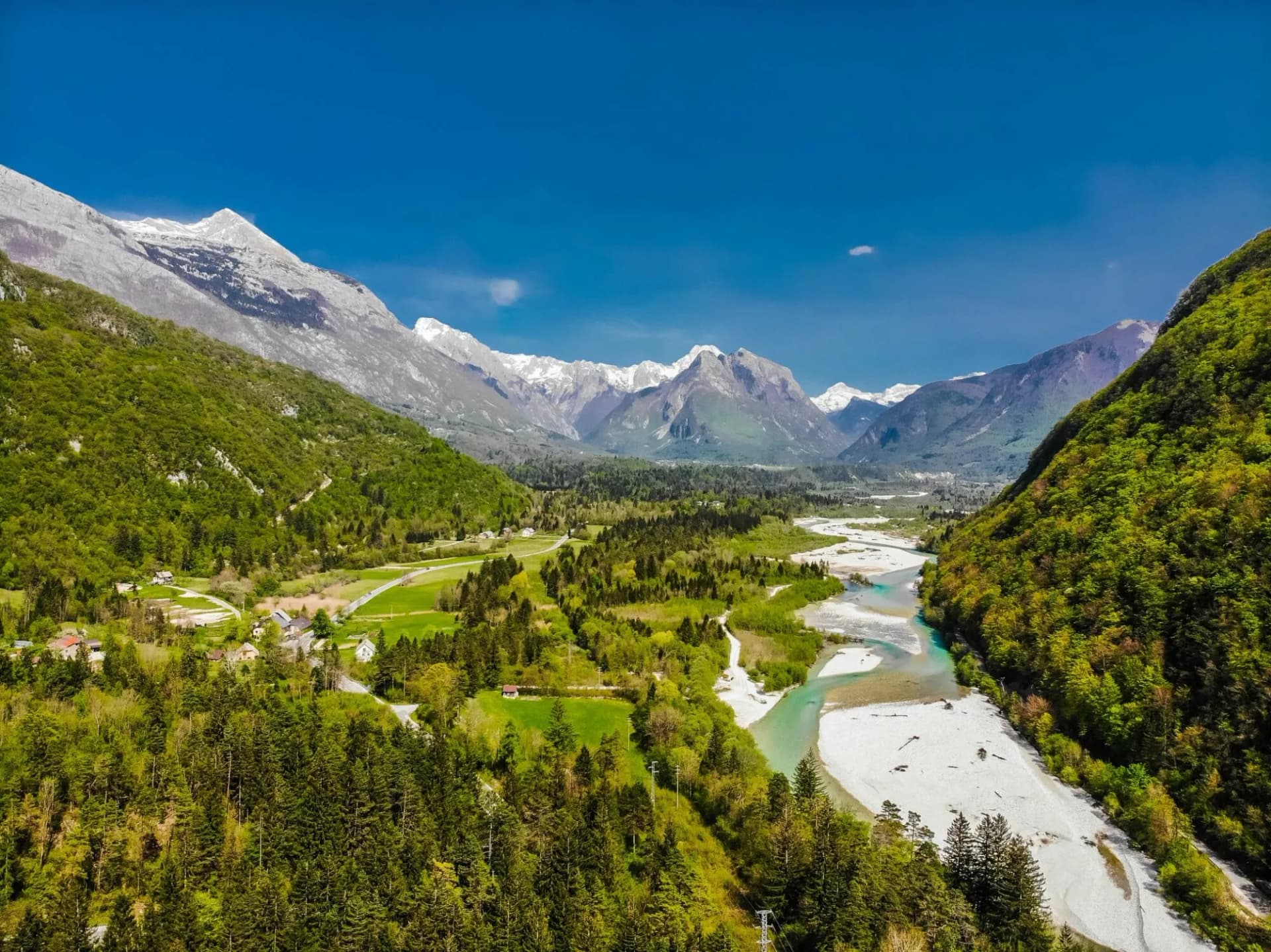 River valley with turquoise water, green forests, and snow-capped mountains under a blue sky.